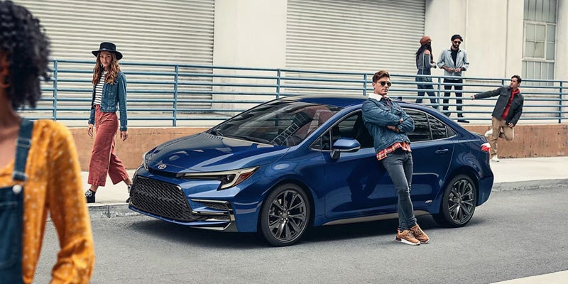 A man leans against a blue toyota Corolla on a city street while pedestrians walk nearby.